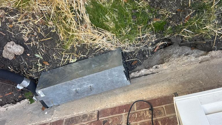Galvanized one-way wildlife trap positioned over an exterior foundation gap during a residential pest control service in The Colony, Texas.