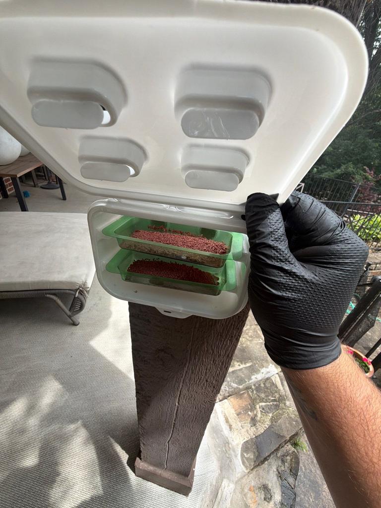 A technician wearing gloves services an exterior fly bait station mounted on a patio post at a residential property in McKinney, Texas.