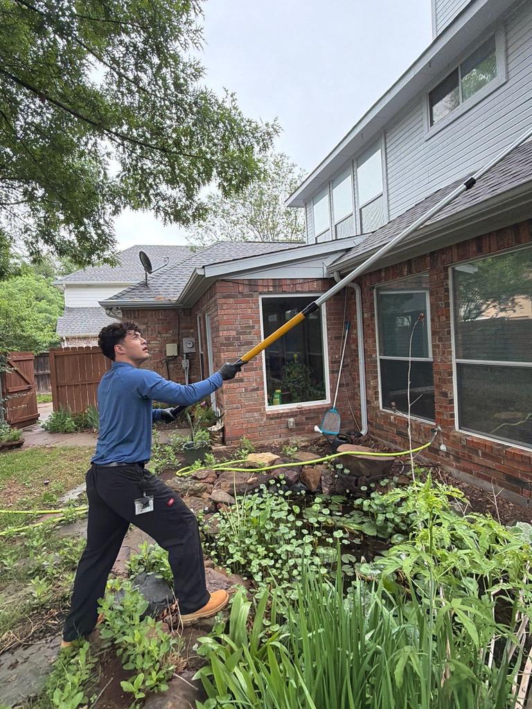 Technician uses an extension pole to remove spider webs from upper eaves above a backyard pond at a brick home in McKinney, Texas.