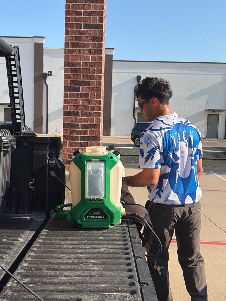 A pest control technician in a blue and white beetle print shirt stands at the open tailgate of a truck, preparing a green FlowZone Typhoon backpack sprayer. 3 A pest control technician stands at the open tailgate of a truck, preparing a green FlowZone Typhoon backpack sprayer on the truck bed.