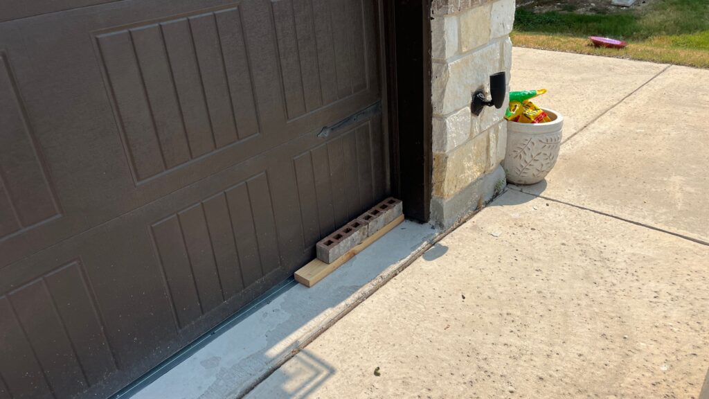 A cinder block and a piece of wood placed to block a gap at the bottom corner of a brown garage door frame on a concrete driveway.
