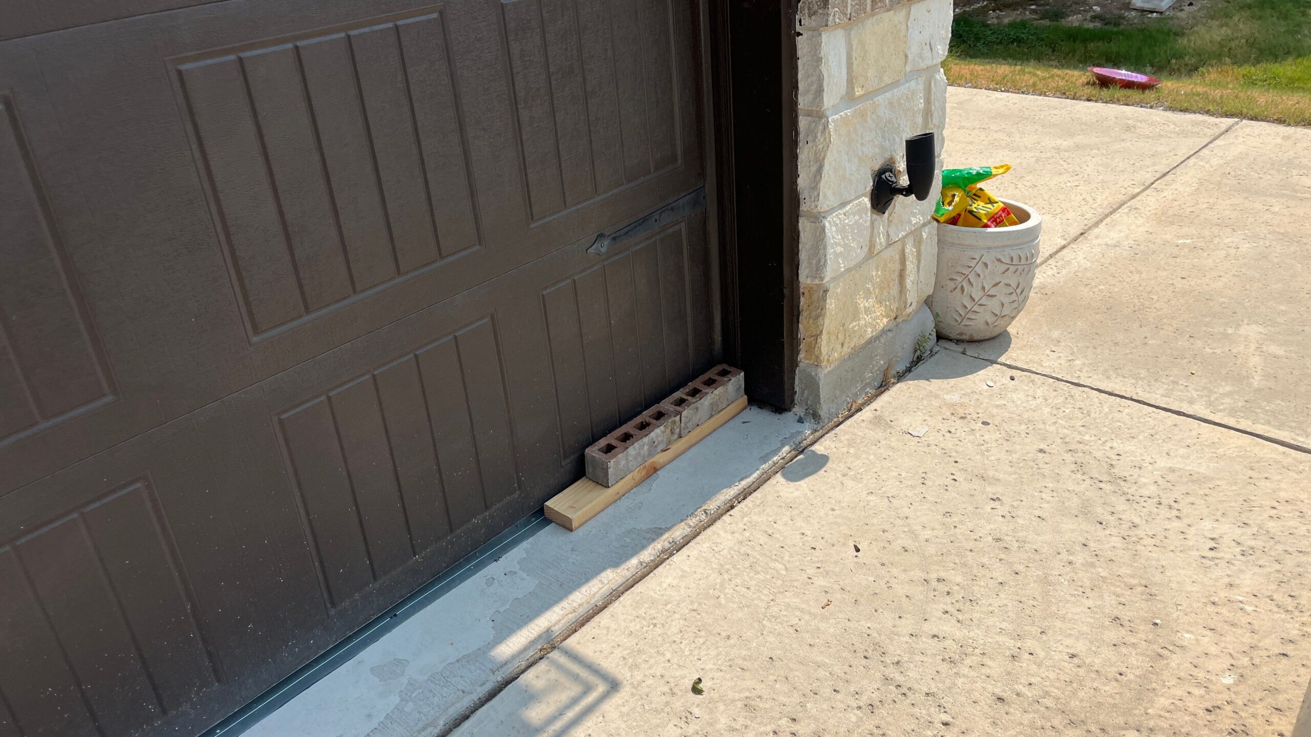 A cinder block and a piece of wood placed to block a gap at the bottom corner of a brown garage door frame on a concrete driveway.