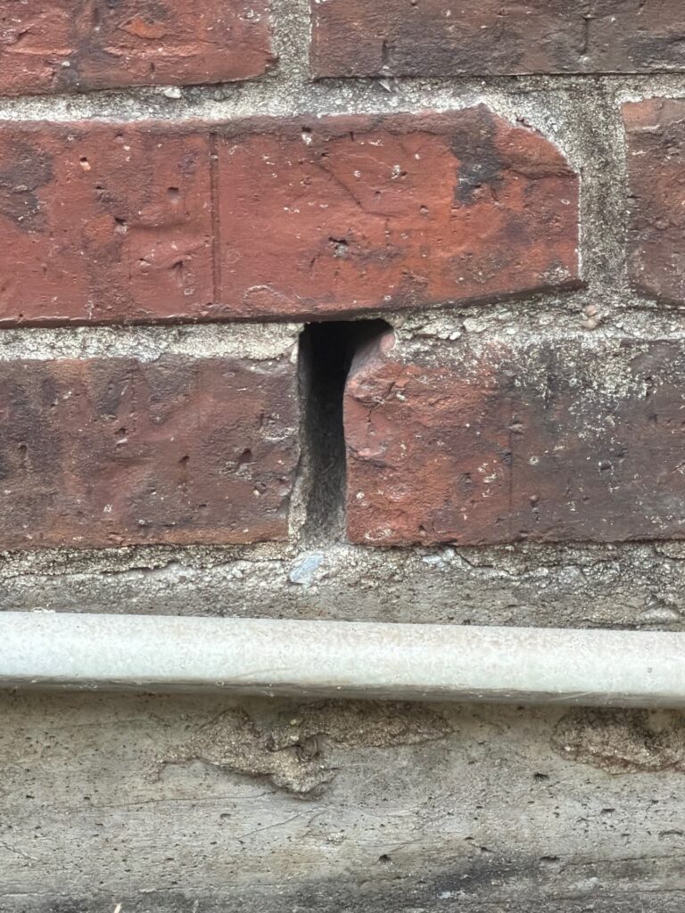 Open Weep Hole in Brick Near Foundation in Allen 3 Close up of an open weep hole gap in a red brick exterior wall just above a pipe near the foundation.