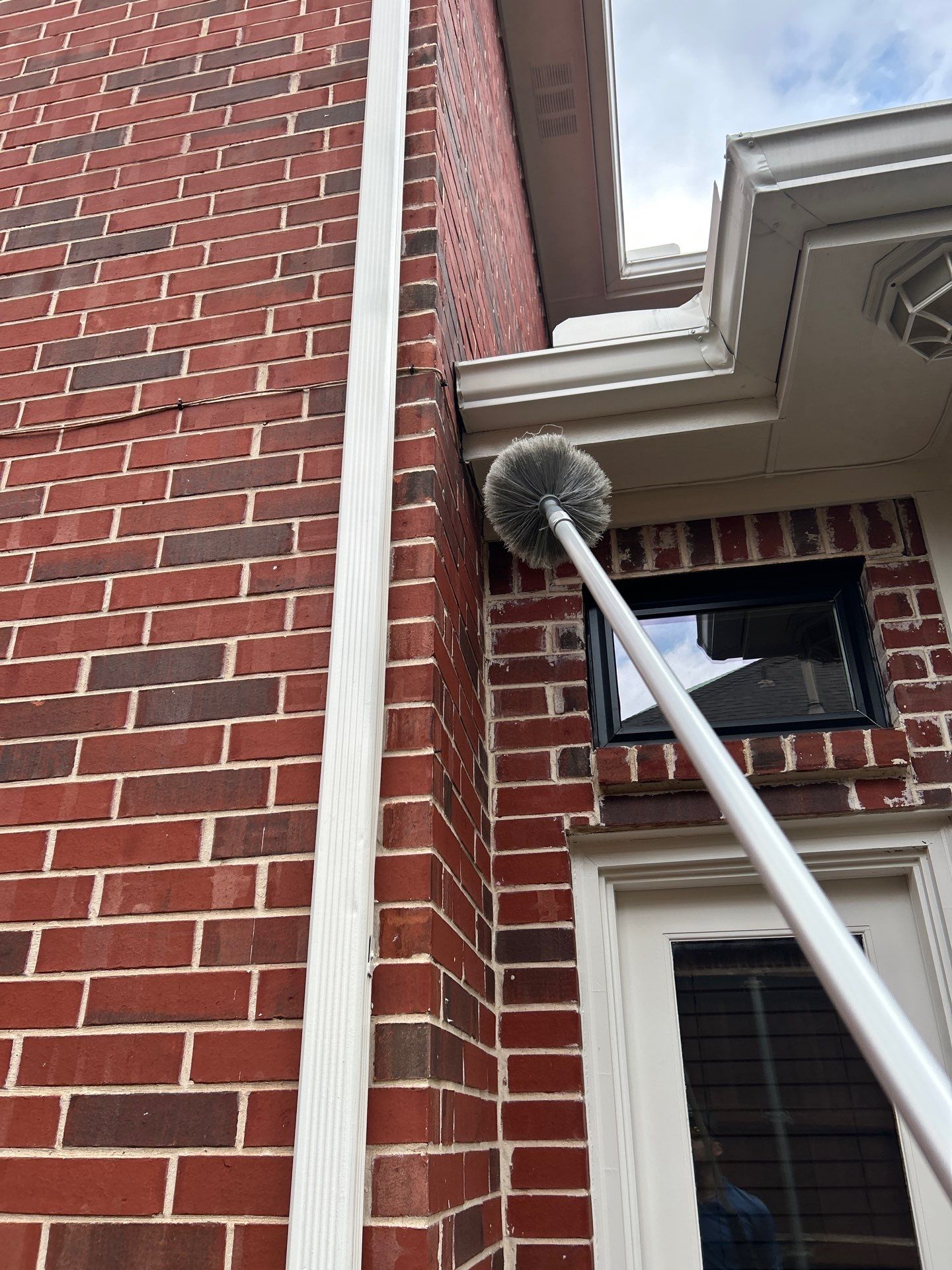 Extension duster pole with a round gray brush reaching up to clear spider webs under a white gutter and soffit on a red brick house, with a window and white back door below.