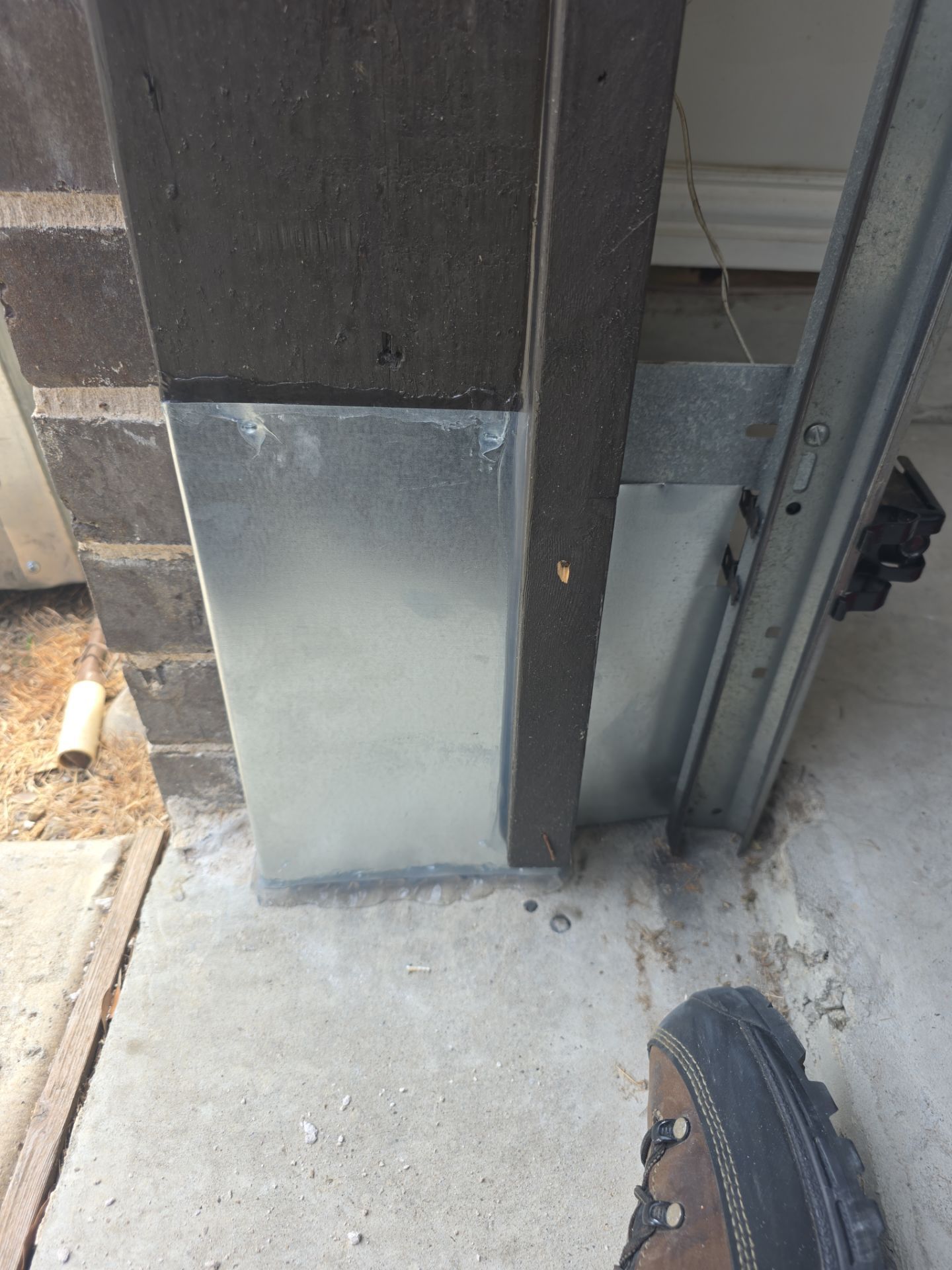 Galvanized metal flashing wrapped around the base of a dark wooden garage door frame, sealed with clear caulk along the top edge and at the concrete floor, with a second metal panel behind the garage door track.