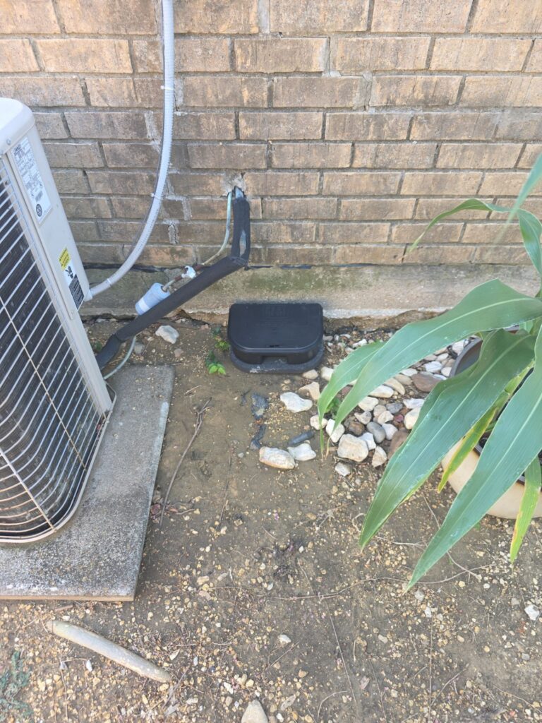 Rodent Bait Station Beside AC Unit in Carrollton 3 A black locking rodent bait station sits on the dirt against a brick wall foundation, between an outdoor AC condenser unit with insulated refrigerant lines and a potted plant on gravel.