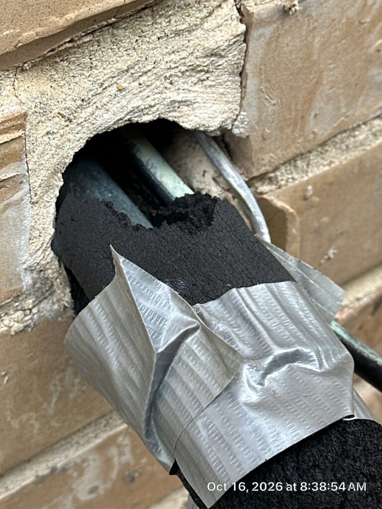 Pest Exclusion & Rodent Proofing 20 Close-up of a large unsealed hole in a tan brick exterior wall where copper AC refrigerant lines wrapped in black foam insulation and silver duct tape enter the house, with open gaps around the pipes.