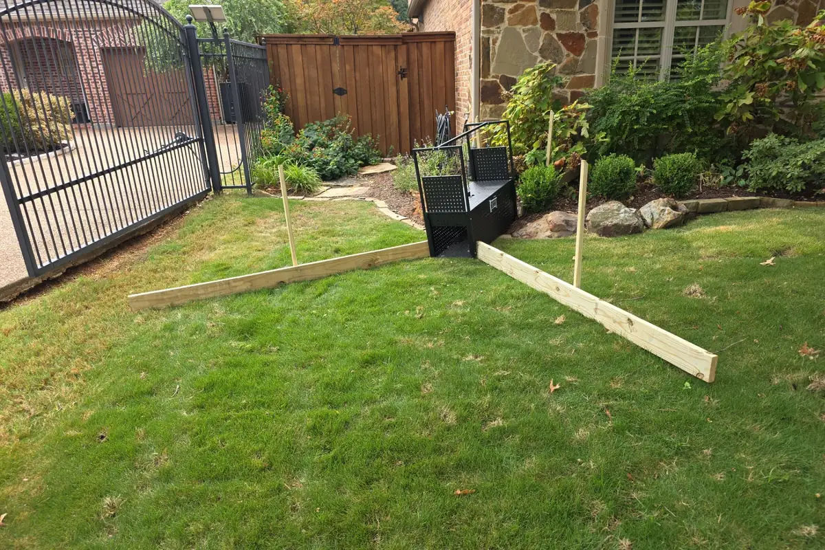 What Is Digging Up My Flower Bed at Night? 14 Black wildlife cage trap at the edge of a front flower bed, with two staked boards on the lawn angled in a V toward its open door